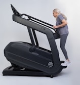 An older adult woman with long gray hair exercises on a large, dark-colored treadmill, dressed in gray activewear and white sneakers.