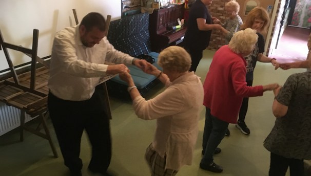 A group of elderly people are engaging in a dance activity in a room. Some chairs are stacked on the side, and a piano is visible in the background. The atmosphere appears lively and social.
