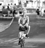 An elderly person is riding a bicycle on a paved path. They appear relaxed, surrounded by several people standing and walking in the background near a railing beside a body of water. The pathway and trees create a serene atmosphere.