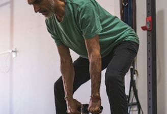 An older man is performing a strength training exercise in a gym. He is wearing a green shirt and dark pants, holding a weight with both hands. The background includes gym equipment such as weight plates and resistance bands.