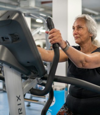 An elderly person is exercising on a stationary bike in a gym setting. The environment is filled with various gym equipment and modern fitness machines. The person appears focused and determined, wearing casual workout attire.