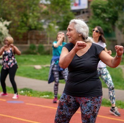 A group of people is participating in an outdoor exercise class on a track. The focus is on an older woman with gray hair wearing a black shirt and colorful leggings, enthusiastically performing workout movements. In the background, several other women are similarly dressed in activewear, exercising with determination. The setting appears to be in a park with lush green trees and bushes.