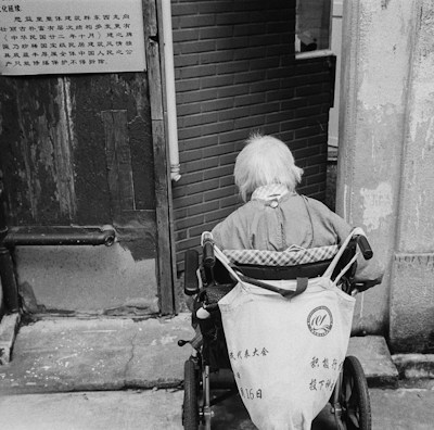 An elderly person with white hair sits in a wheelchair facing away from the camera. They are positioned near a worn doorway with textured walls and a brick interior visible beyond. A sign in an East Asian script is attached to the door nearby.
