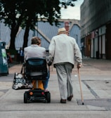 Two elderly individuals are moving along a paved path in an urban setting. One person is using a mobility scooter and the other is walking with a cane. They are both dressed in light-colored clothing and surrounded by buildings and trees.