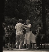 A group of elderly people is gathered in a park, engaging in social interaction. Some are dancing while others watch and converse. The setting is shaded by trees, creating a peaceful and intimate atmosphere.