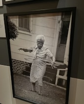 An elderly woman is using a hula hoop in a yard. She is wearing a patterned dress and appears focused on the activity. The background shows a house with a white exterior and a brick foundation. The photo is in black and white, adding a nostalgic feel.