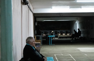 An elderly person sits on a bench in a dimly lit indoor space with marked flooring. In the background, more people are visible, seated and engaged in different activities. The setting suggests a community or recreational area, possibly a basement or hallway with a slightly worn appearance.