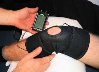 A person is applying a black knee brace to another person's leg, which is resting on a pillow. The person is also holding a handheld electronic device with a small screen displaying some controls and information, likely related to therapy or treatment.