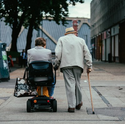 Two elderly individuals are moving along a paved path in an urban setting. One person is using a mobility scooter and the other is walking with a cane. They are both dressed in light-colored clothing and surrounded by buildings and trees.