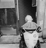 An elderly person with white hair sits in a wheelchair facing away from the camera. They are positioned near a worn doorway with textured walls and a brick interior visible beyond. A sign in an East Asian script is attached to the door nearby.