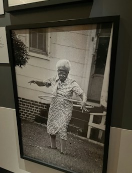 An elderly woman is using a hula hoop in a yard. She is wearing a patterned dress and appears focused on the activity. The background shows a house with a white exterior and a brick foundation. The photo is in black and white, adding a nostalgic feel.