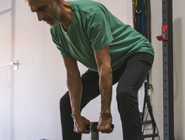 An older man is performing a strength training exercise in a gym. He is wearing a green shirt and dark pants, holding a weight with both hands. The background includes gym equipment such as weight plates and resistance bands.
