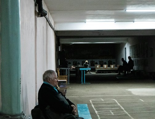 An elderly person sits on a bench in a dimly lit indoor space with marked flooring. In the background, more people are visible, seated and engaged in different activities. The setting suggests a community or recreational area, possibly a basement or hallway with a slightly worn appearance.