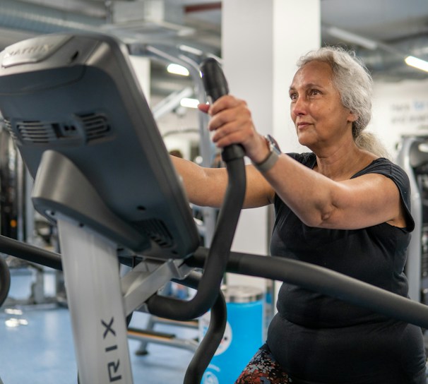 An elderly person is exercising on a stationary bike in a gym setting. The environment is filled with various gym equipment and modern fitness machines. The person appears focused and determined, wearing casual workout attire.