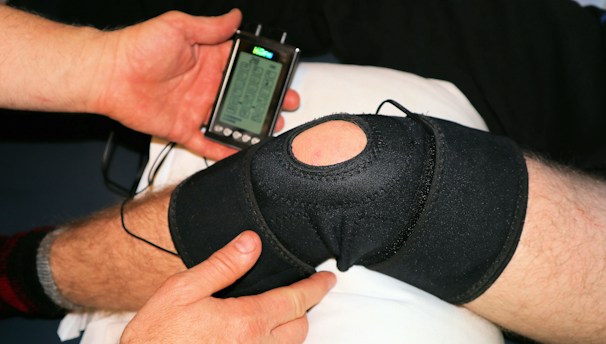 A person is applying a black knee brace to another person's leg, which is resting on a pillow. The person is also holding a handheld electronic device with a small screen displaying some controls and information, likely related to therapy or treatment.
