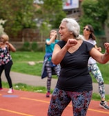 A group of people is participating in an outdoor exercise class on a track. The focus is on an older woman with gray hair wearing a black shirt and colorful leggings, enthusiastically performing workout movements. In the background, several other women are similarly dressed in activewear, exercising with determination. The setting appears to be in a park with lush green trees and bushes.