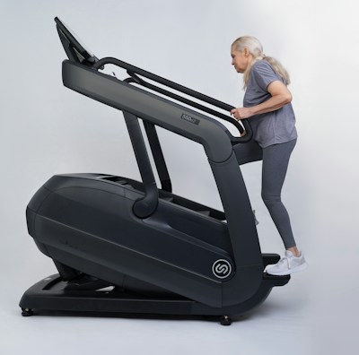 An older adult woman with long gray hair exercises on a large, dark-colored treadmill, dressed in gray activewear and white sneakers.