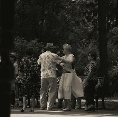 A group of elderly people is gathered in a park, engaging in social interaction. Some are dancing while others watch and converse. The setting is shaded by trees, creating a peaceful and intimate atmosphere.