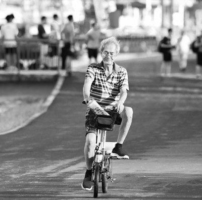An elderly person is riding a bicycle on a paved path. They appear relaxed, surrounded by several people standing and walking in the background near a railing beside a body of water. The pathway and trees create a serene atmosphere.